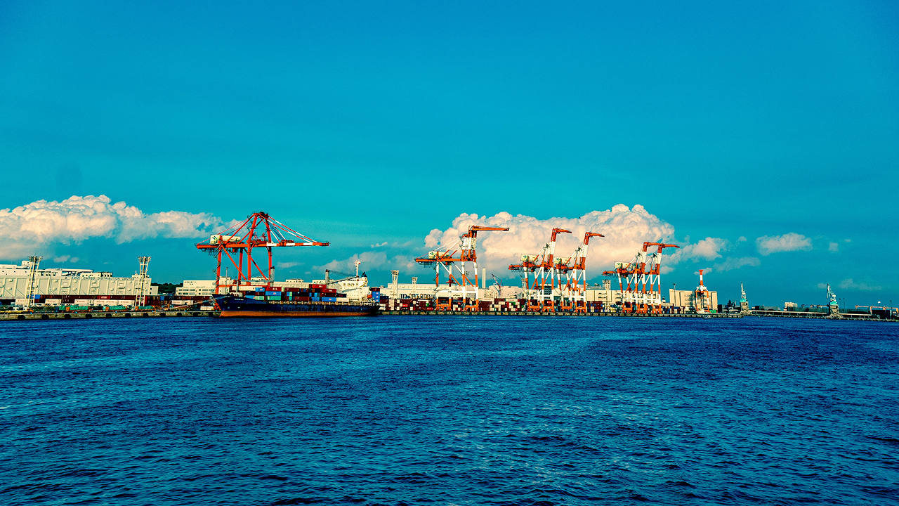 Container vessel berthed at a port terminal with quay cranes, container yard, and stacked shipping containers.